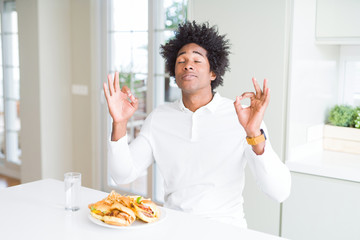 Fototapeta na wymiar African American hungry man eating hamburger for lunch relax and smiling with eyes closed doing meditation gesture with fingers. Yoga concept.