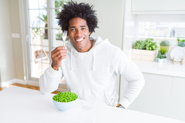 African American man eating fresh green peas at home with a happy face standing and smiling with a...