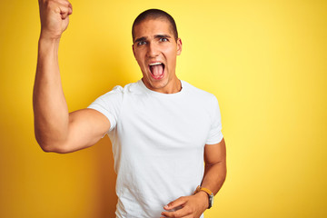Young caucasian man wearing casual white t-shirt over yellow isolated background angry and mad raising fist frustrated and furious while shouting with anger. Rage and aggressive concept.