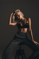 Fototapeta na wymiar Fashion young beauty woman with blonde hair in lingerie and classic trousers posing on chair on black background in room