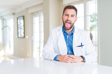Naklejka na ściany i meble Handsome doctor man wearing medical coat at the clinic sticking tongue out happy with funny expression. Emotion concept.