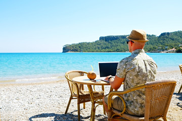 Fit travel blogger sitting at beach bar writing article on white laptop. Freelance remote work concept. Self employed man coding, wearing typical tourist shirt and hat. Copy space, sea view background