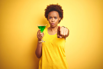 Young african american woman with afro hair drinking a cocktail over yellow isolated background pointing with finger to the camera and to you, hand sign, positive and confident gesture from the front