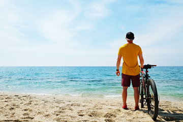 Young man with fit body riding the mtb mountain bike on sandy beach with beautiful azure water sea view. Muscle male wearing bright yellow t-shirt cycling on ocean shore. Close up, copy space.