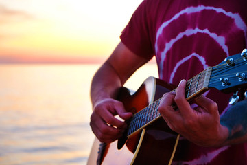 Young man wearing purple tie dye t-shirt playing dreadnought parlor acoustic guitar on beach at...
