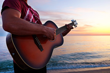 Young man wearing purple tie dye t-shirt playing dreadnought parlor acoustic guitar on beach at...