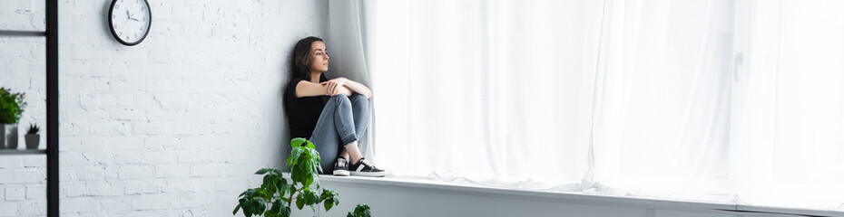 panoramic shot of depressed young woman sitting on window sill with crossed arms and looking away - obrazy, fototapety, plakaty