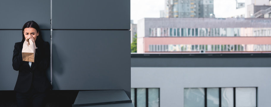 panoramic shot of scared businesswoman standing on rooftop and breathing into paper bag while suffering from panic attack