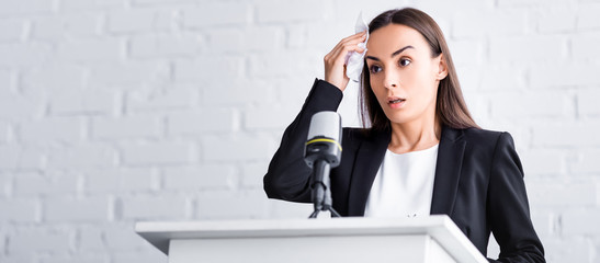 panoramic shot of worried lecturer suffering from fear of public speaking holding napkin near forehead while standing on podium tribune