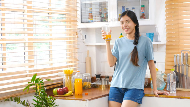 Young beautiful asian woman drinking orange juice and smiling while standing by window in kitchen background, peolpe and healthy lifestyles