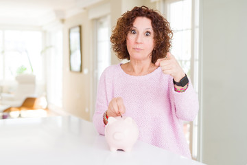 Senior woman saving money putting a coin inside piggy bank pointing with finger to the camera and to you, hand sign, positive and confident gesture from the front