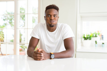 Handsome african american man on white table at home puffing cheeks with funny face. Mouth inflated with air, crazy expression.