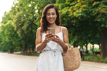 Portrait of happy woman smiling and holding cellphone while walking through green boulevard