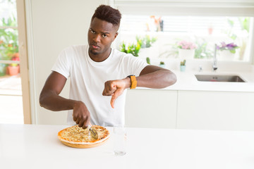 African american man eating cheese pizza at home with angry face, negative sign showing dislike...
