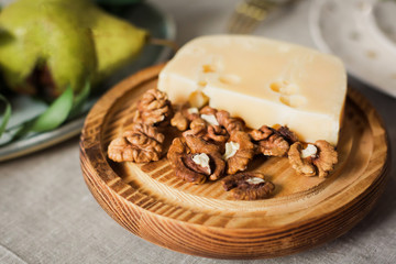A piece of tasty hard milk cheese in a hole lies on a wooden board with walnut kernels against a served table with a ripe big pear