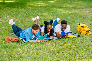 Front view of three happy multiethnic students studying reading notes together lying on the grass...
