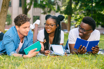 Front view of three happy multiethnic students lying, talking, joking together together on green...