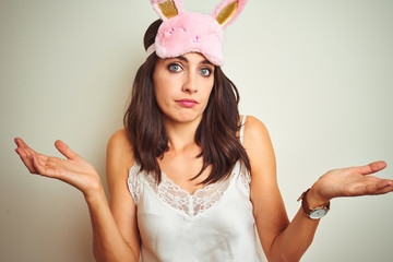 Young beautiful woman wearing pajama and mask standing over white isolated background clueless and...