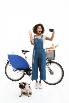 Image of joyful african american woman taking selfie photo on smartphone while standing with her pug and bicycle