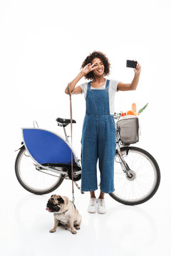 Image of beautiful african american woman taking selfie photo on smartphone while standing with her pug and bicycle