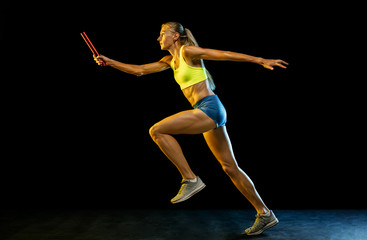 Professional female relay racer training isolated on black studio background in neon light. Woman in sportsuit practicing in running. Healthy lifestyle, sport, workout, motion, action concept.