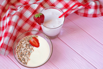 A dietary breakfast of a glass of strawberry yogurt, a deep plate of muesli with strawberries and a plate of sour cream are set on a delicate pink wooden background with a red and white check cotton