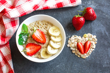 Porridge with strawberries and banana slices in a white plate on a dark concrete background. Decoration of dry oatmeal and cloves of strawberries and cotton towels in a red
