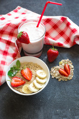 Porridge with strawberries and banana slices in a white plate, yogurt in a glass stand on a dark concrete background. Decoration of dry oatmeal and strawberry cloves.