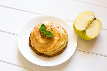 On a white plate lies a cake of apple charlotte with mint leaves close-up on a white wooden background. Nearby lies a half of fresh green apple.
