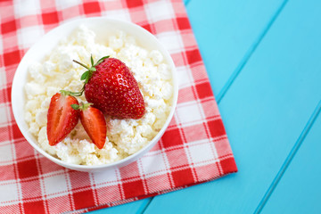 Fresh tasty granular cottage cheese with strawberries in a white deep plate on a blue wooden background and beside a cotton towel in a red-white cage