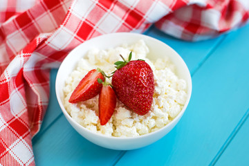 Fresh tasty granular cottage cheese with strawberries in a white deep plate on a blue wooden background and beside a cotton towel in a red-white cage