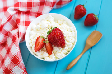 Fresh tasty granular cottage cheese with strawberries in a white deep plate on a blue wooden background and beside a cotton towel in a red-white cage, strawberries and a wooden spoon