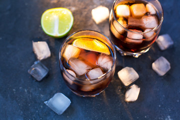  Two glasses with cocktails: rum cola and cola whiskey stand on a gray blue concrete textured background and ice cubes and chopped lime lie around