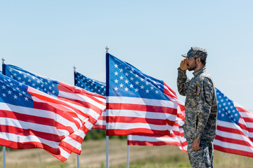 Man in military uniform and cap standing and giving salute near American flags