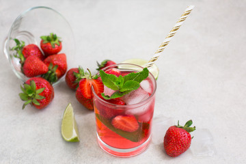 On a light background there is a strawberry cocktail in a glass with ice and mint, a bowl with strawberries and lime slices