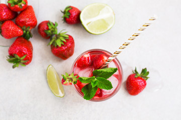 On a light gray background there is a strawberry cocktail in a glass with ice and mint, an inverted bowl with strawberries and slices of sliced lime