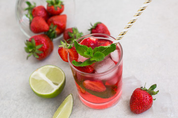 On a light background there is a strawberry cocktail in a glass with ice and mint, a bowl with strawberries and lime slices