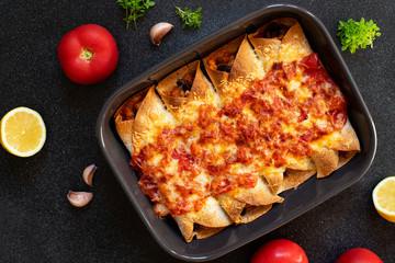 Mexican enchiladas with chicken, vegetables, corn, beans, tomato sauce and cheese. Served in baking tray. Mexican food. Latin American cuisine. Black background, top view, copy space