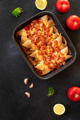 Mexican enchiladas with chicken, vegetables, corn, beans, tomato sauce and cheese. Served in baking tray. Mexican food. Latin American cuisine. Black background, top view, copy space