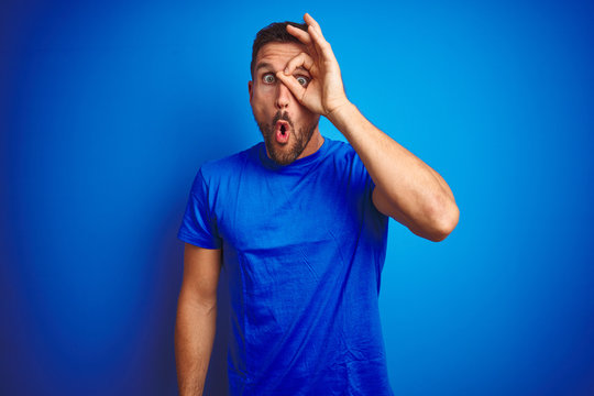 Young handsome man wearing casual t-shirt over blue isolated background doing ok gesture shocked with surprised face, eye looking through fingers. Unbelieving expression.