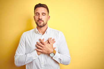 Young handsome man wearing elegant white shirt over yellow isolated background smiling with hands on chest with closed eyes and grateful gesture on face. Health concept.