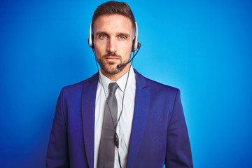 Young handsome operator man wearing call center headset over blue isolated background with serious expression on face. Simple and natural looking at the camera.