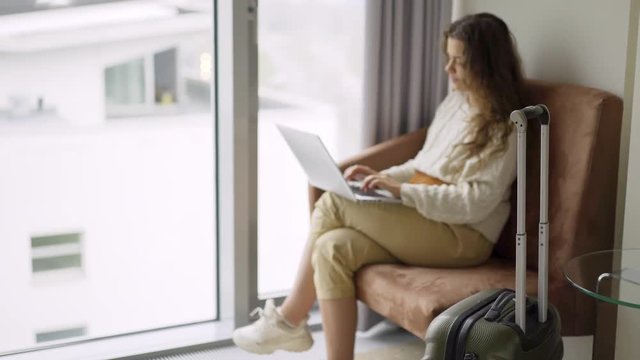 Young woman arrived modern hotel suite. Side view female sitting in armchair near full length window with laptop. Defocused girl relaxing after journey, working on computer and enjoying view.