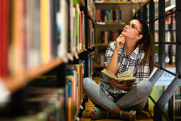 Young female student study in the library reading book while sitting near bookshelf.