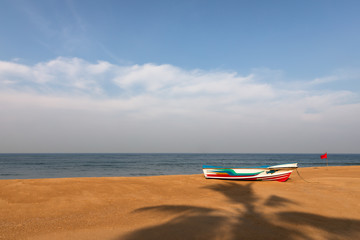 A colorful boat on the sand beach of the Indian Ocean at Mount Lavinia, Sri Lanka with the shadow of a palm tree and a red flag.