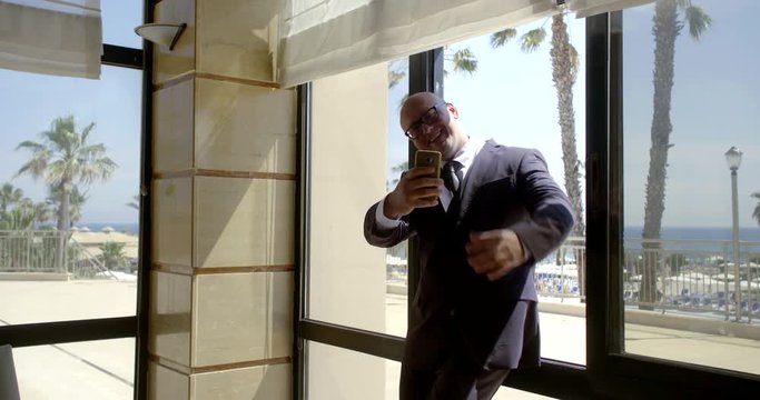 Bald business man in glasses and a classic suit comes to the panoramic window at the reception, photographed on the phone, takes a selfie and smiles near the Windows