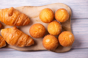 View from above of cupcakes and croissants prepared for breakfast on wooden board on white wooden table.