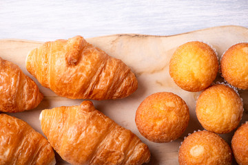 View from above of cupcakes and croissants prepared for breakfast on wooden board on white wooden table.