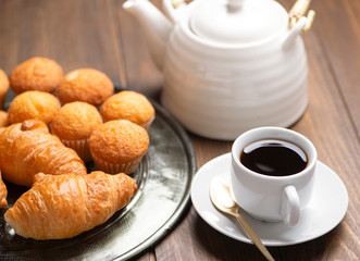 Close-up of cupcakes and croissants on metal tray next to cup of coffee and coffee pot. Breakfast.