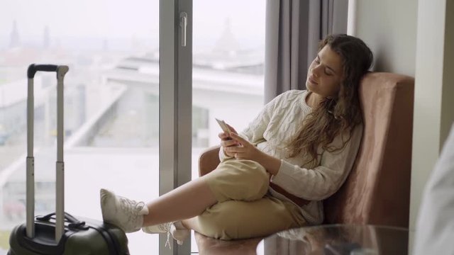 Young woman arrived modern hotel suite. Side view girl sitting in armchair near full length window with phone. She relaxing after journey, browsing on smartphone and enjoying view above city